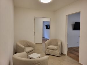 Raum mit drei grauen Sesseln, einem kleinen weißen Tisch und zwei Türen die in Nebenräume führen